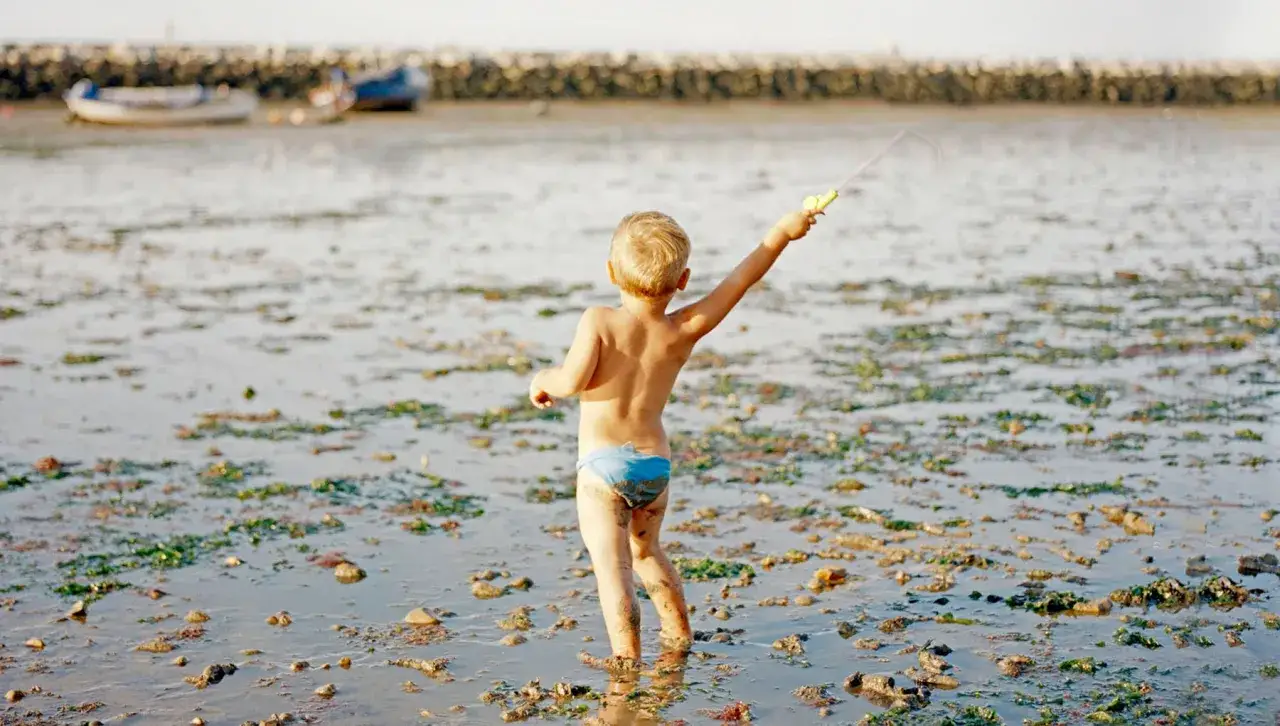 Bambino con canna da pesca in mano, in piedi nell'acqua bassa, con alghe e rocce. Un'immagine che evoca la spensieratezza dell'infanzia, come quelle di Laura Pannack.