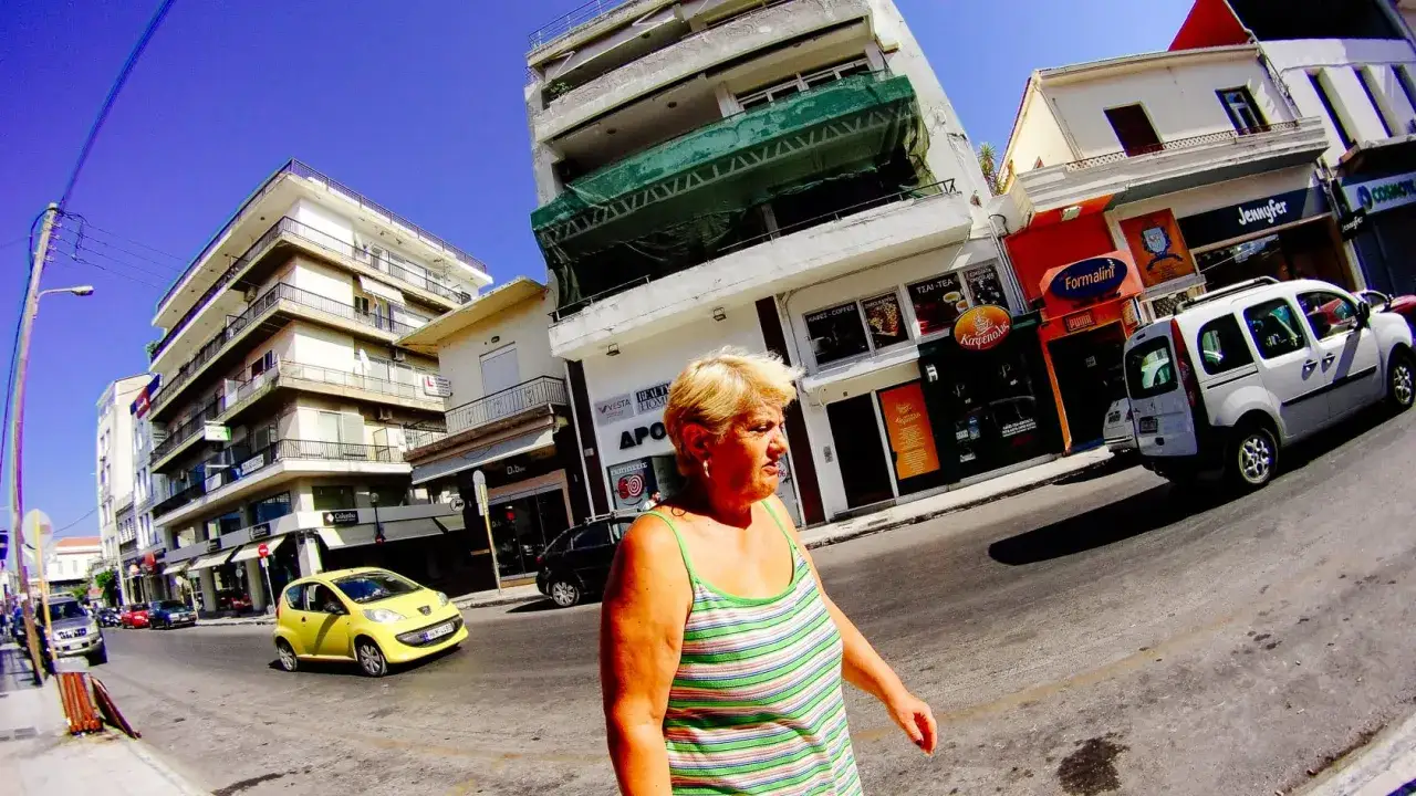 Donna cammina per strada, con edifici e negozi sullo sfondo. Una piccola auto gialla, un furgone bianco e altre auto sono visibili. L'immagine è stata catturata con una GoPro Ace Pro 2.