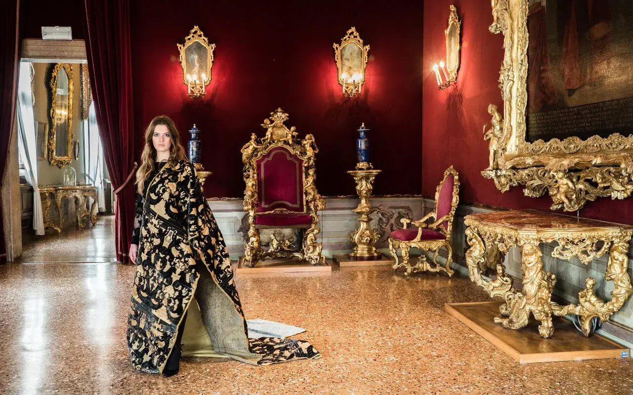 Donna in abito sontuoso con tessitura Bevilacqua visita una sala dorata con trono e specchi antichi.