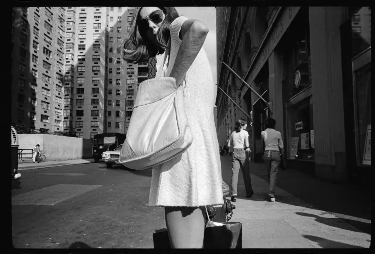 Una donna con borsa a tracolla, in primo piano, cammina per strada. Mark Cohen photography cattura la vita urbana.