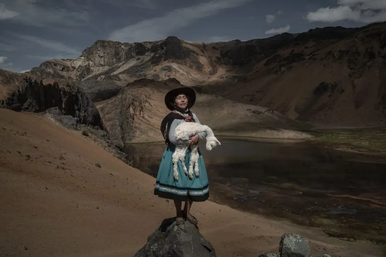 Donna con agnello in braccio, in piedi su una roccia, con montagne e lago sullo sfondo. Alessandro Cinque cattura la serenità.