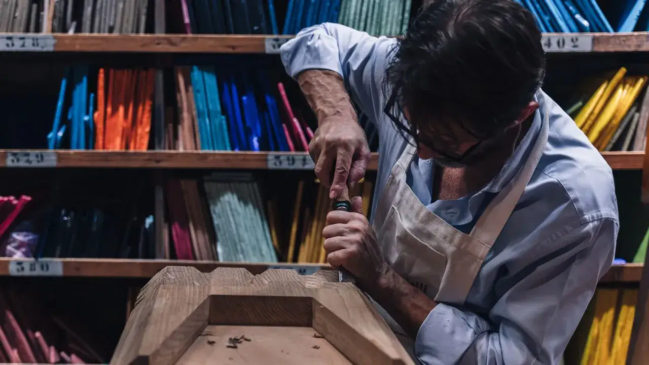 Artigiano al lavoro su un pezzo di legno, con scaffali pieni di campioni colorati alle spalle, un omaggio alla fondazione Cologni.