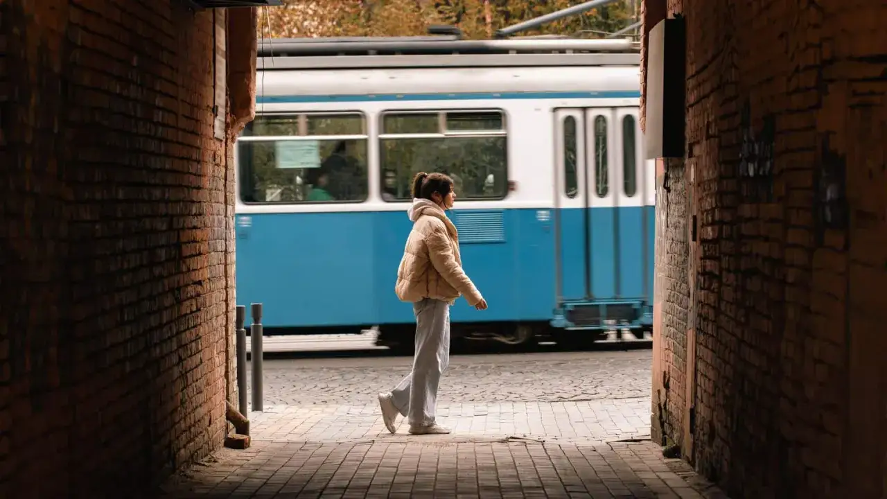 Una giovane donna cammina lungo un vicolo di mattoni, con un tram blu che sfreccia sullo sfondo. Una fotografia di viaggio che cattura un momento urbano.