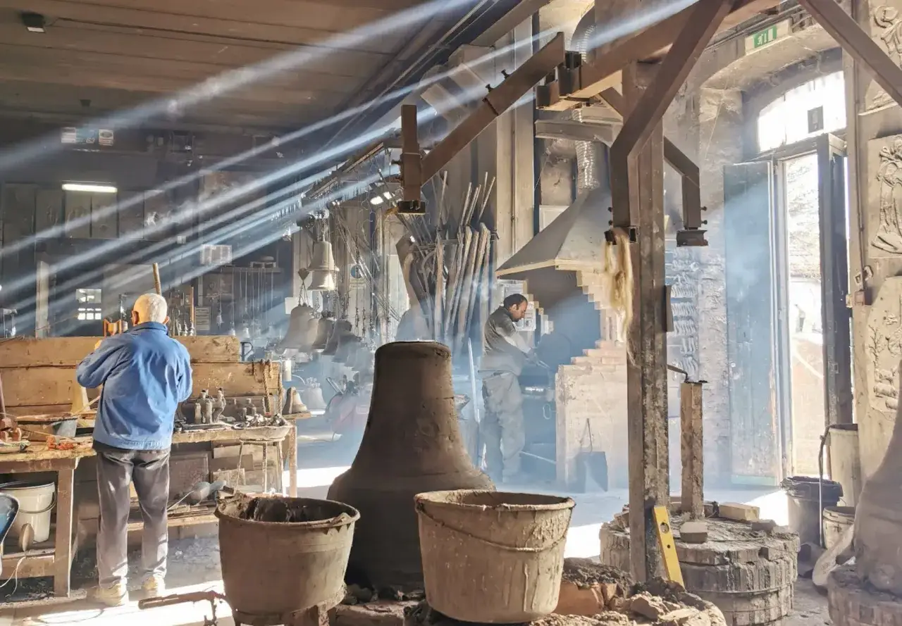 Artigiani al lavoro in una fonderia di Agnone campane. Raggi di sole illuminano il fumo e gli attrezzi, mentre le campane attendono di prendere forma.